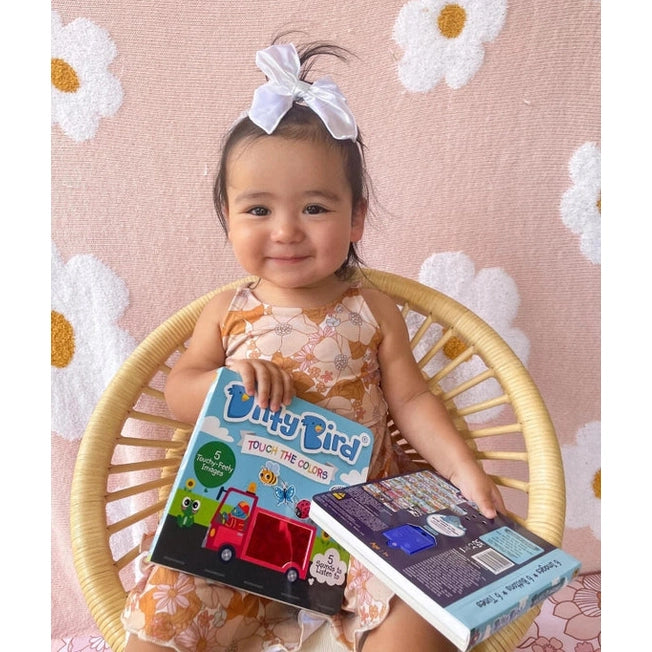 Ditty Bird Sound Book Touch the Colors USB-C Rechargeable: baby girl in a floral dress sits in a wicker chair, holding two colorful Ditty Bird books.