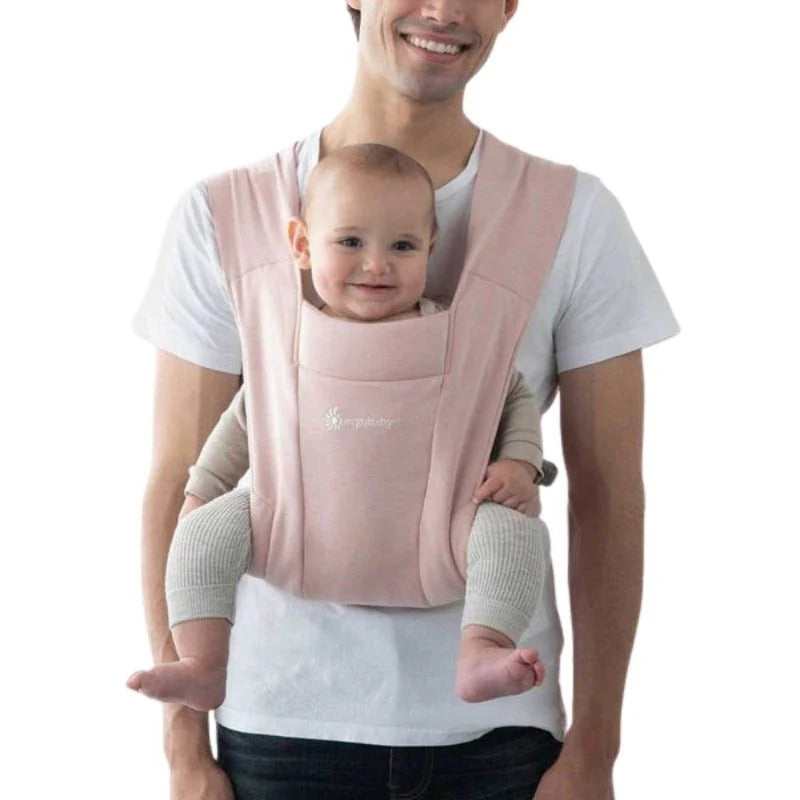 ERGOBABY Embrace Newborn Carrier Blush Pink worn by a parent with a smiling baby secured facing inward.