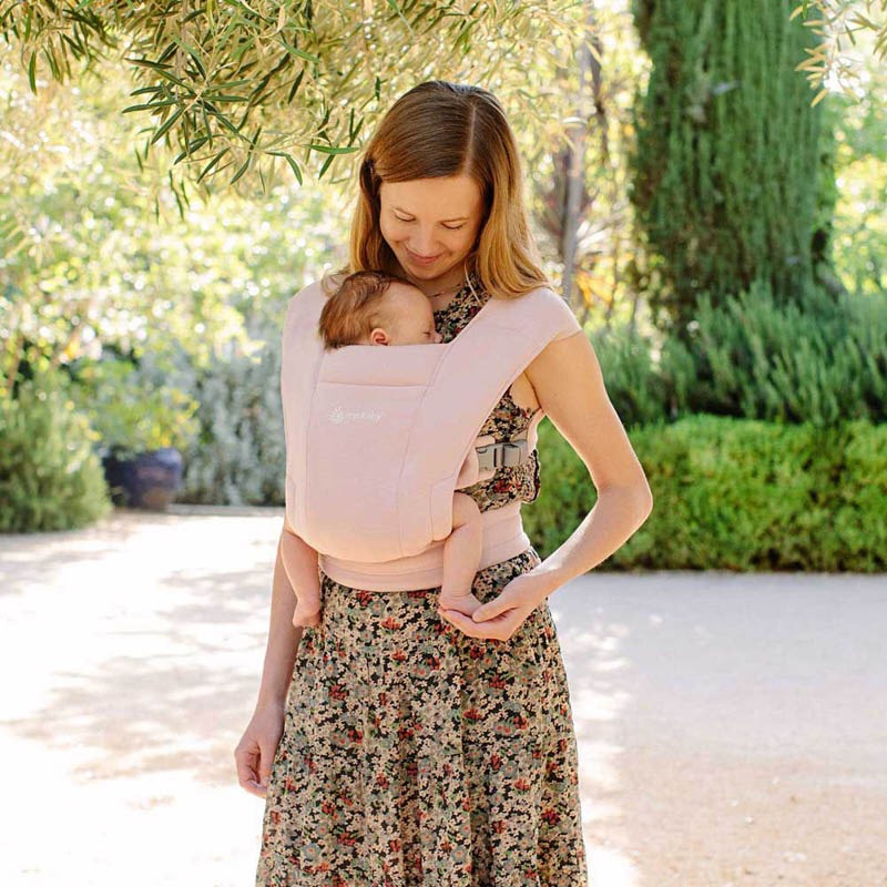 Ergobaby Embrace Newborn Carrier Blush Pink worn by a mom outdoors with baby snug in the soft-structured carrier.