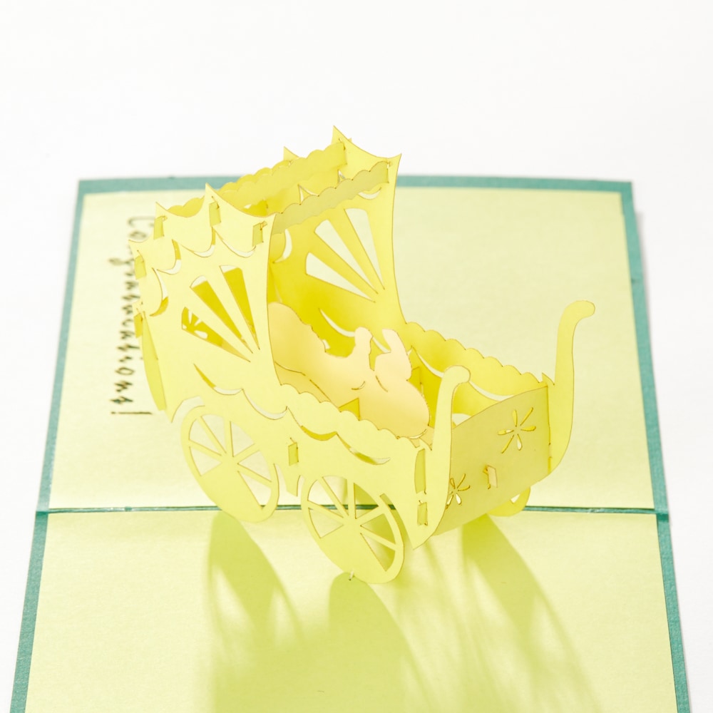 Intricate Greetings Cards Baby Carriage yellow papercut design, opened to reveal delicate wheels and ornate carriage cutouts.