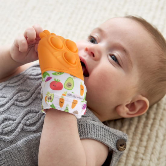 Itzy Ritzy Mitt Pop teething mitten with orange silicone grip; baby gnaws the fruit-printed fabric while lying on a rug.
