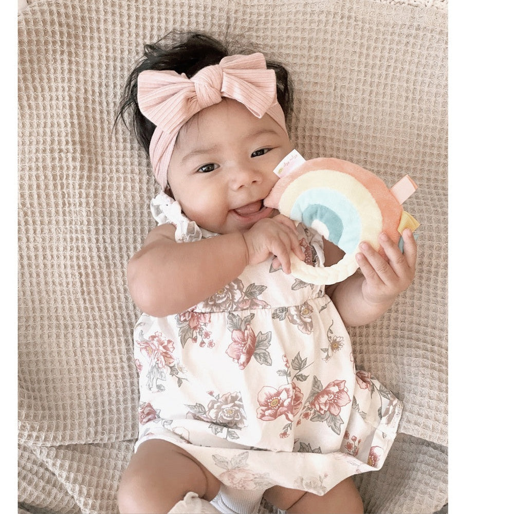 Itzy Ritzy Ritzy Rattle Pal plush rattle with teether; baby girl in a pink headband holds a rainbow arc teether.