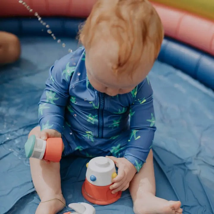 Itzy Ritzy Stack & Squirt bath toy; toddler in a blue swimsuit stacks and squirts colorful cups in an inflatable pool.