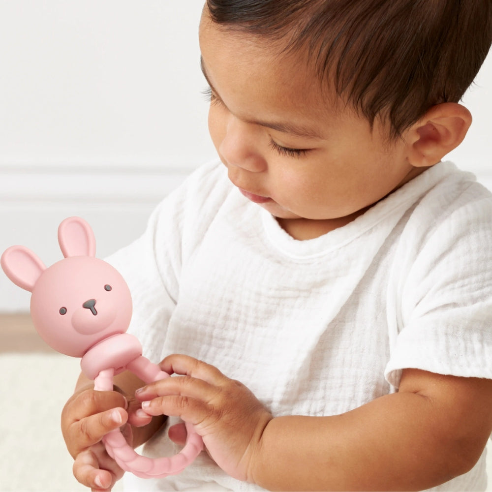 Itzy Ritzy Sweetie Rattle Pink Bunny, a soft pink baby rattle with a loop handle, being held by a curious baby.