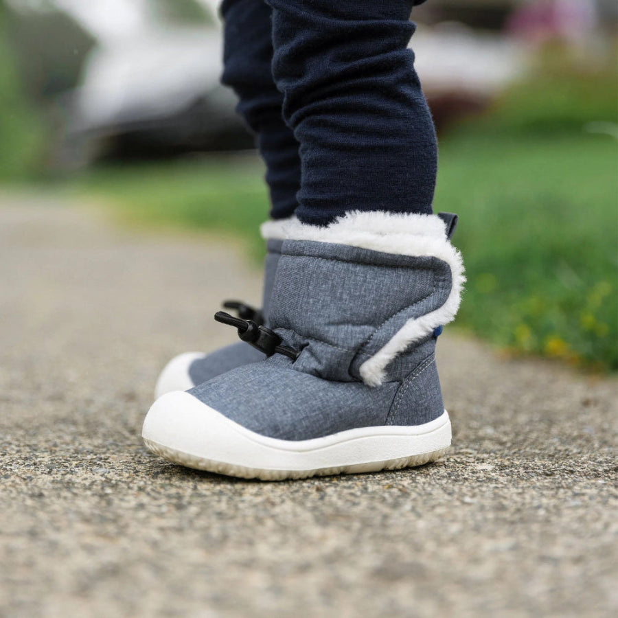 JAN & JUL Baby Winter Boots Bear in grey fabric with plush white lining and white sole; toddler stands on a sidewalk.