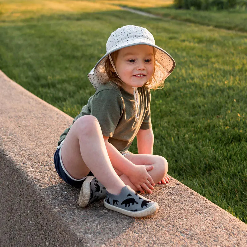 JAN & JUL Cotton Adventure Hat Black Tie-Dye worn by a toddler sitting on a sunlit park curb.