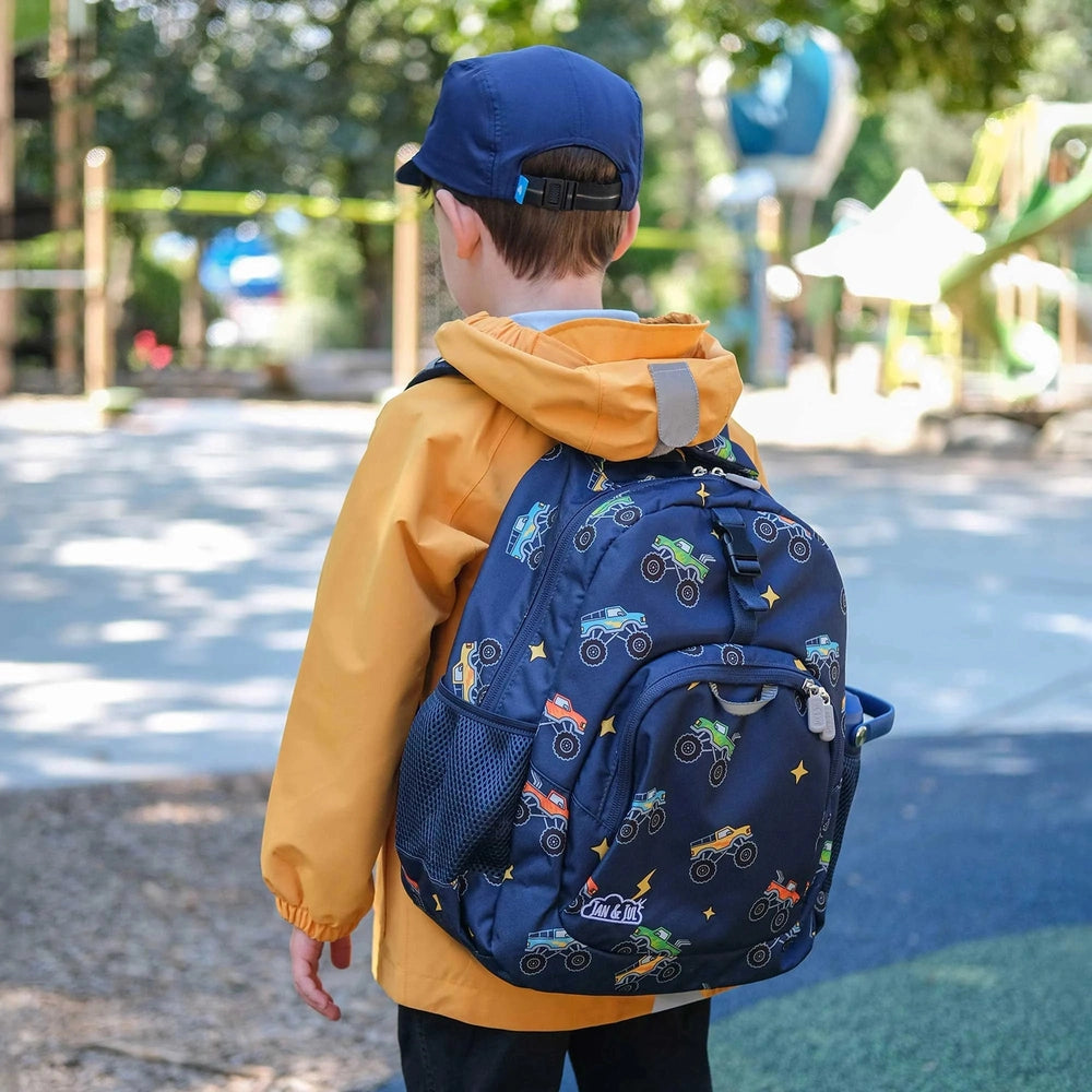 Jan & Jul Kids Backpack navy with colorful monster-truck pattern; child walks in park showing side mesh pocket.