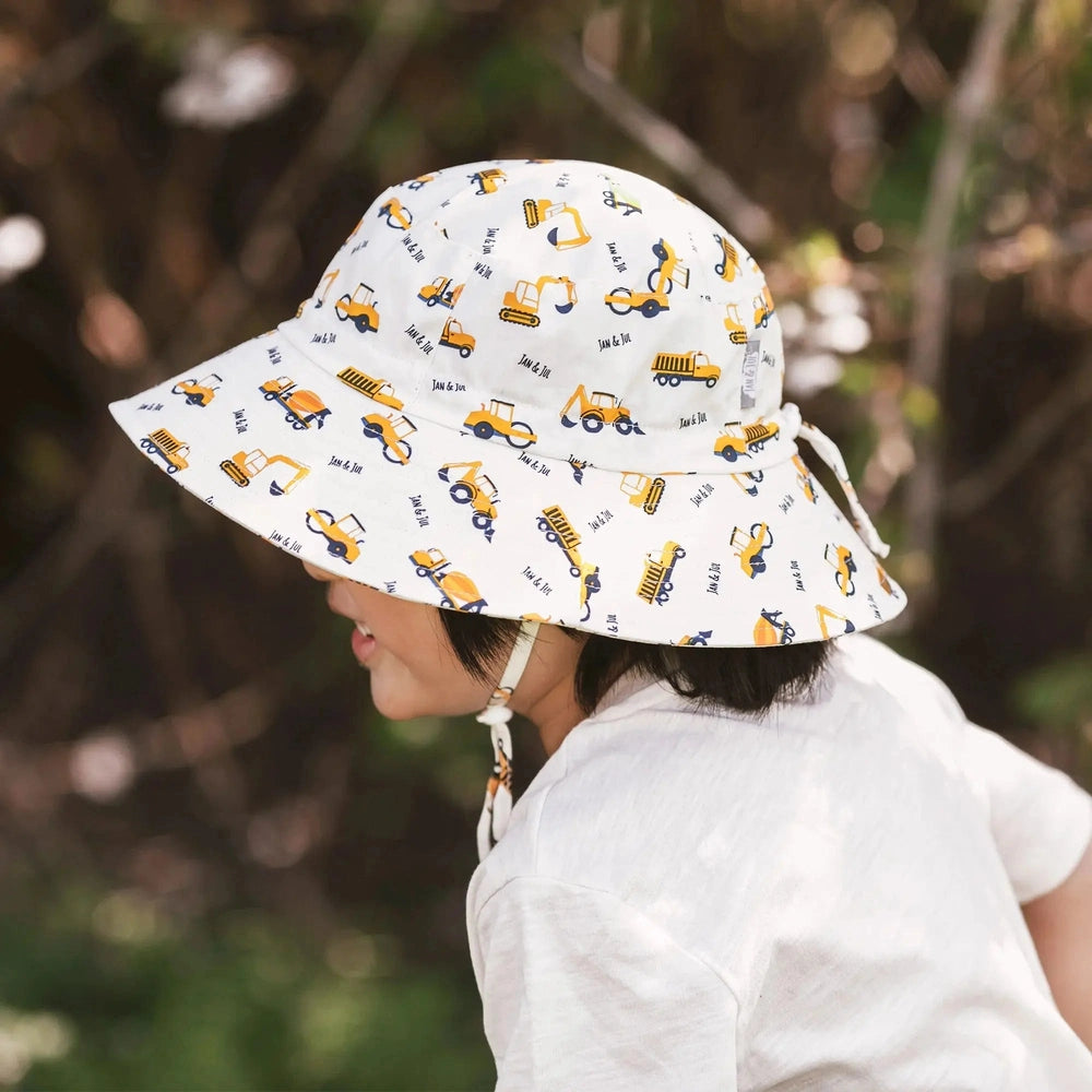 Jan & Jul Kids Cotton Bucket Hat Little Diggers, white fabric with yellow construction vehicles, worn by a toddler.