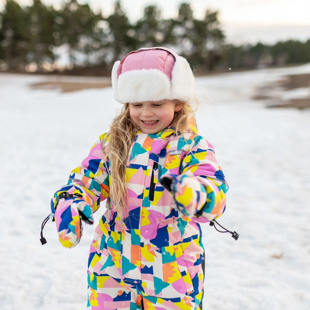 JAN & JUL Kids Insulated Winter Hats Dusty Pink on a smiling girl in a colorful snowsuit, adjusting the hat.