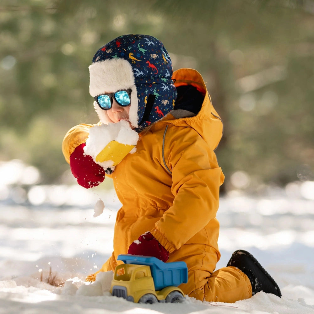 Jan & Jul Kids Insulated Winter Hats Space Dinos in a blue dinosaur print with faux fur trim; child tosses snow.