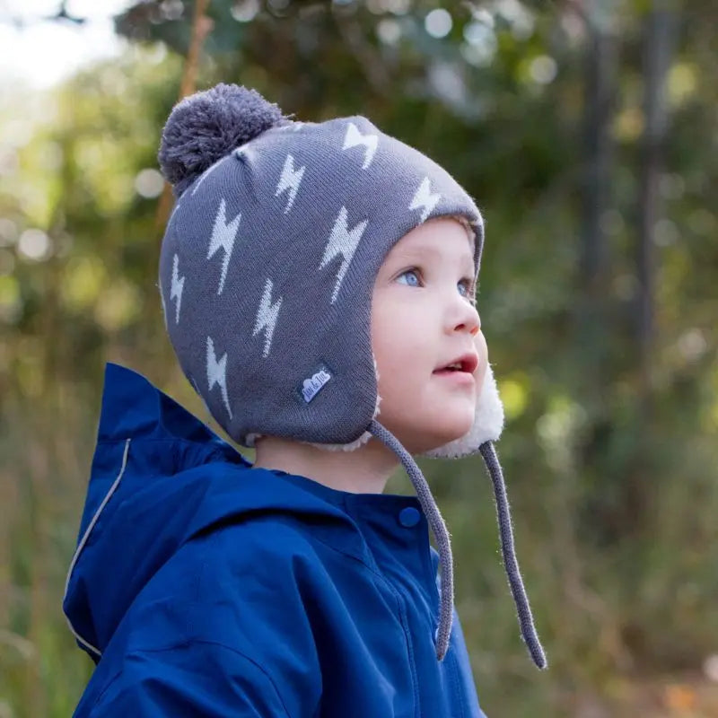 JAN & JUL Kids Knitted Beanie Hat Grey Lightning Bolt with pom-pom, worn by a child outdoors.