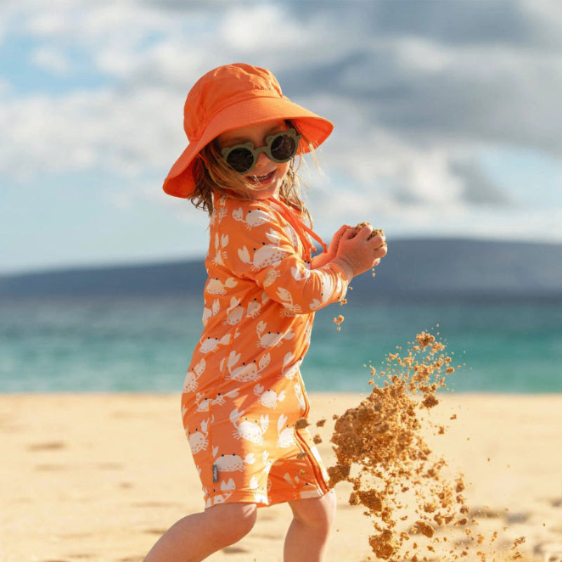 Jan & Jul Kids One Piece UV Sun Suit Crabby Crab orange swimsuit; toddler scoops sand at the sunny beach.