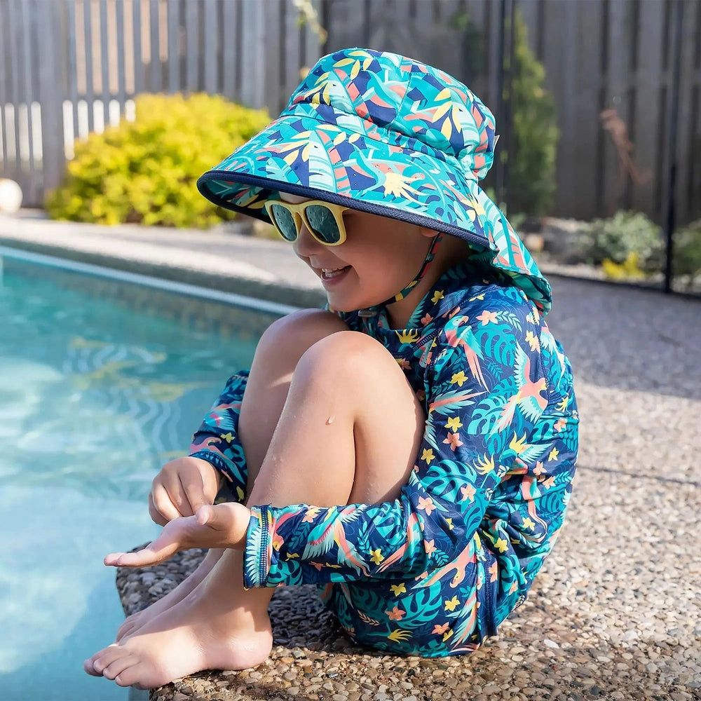 Jan & Jul Kids Water Repellent Adventure Hats Tropical worn by a child sitting by a pool in teal tropical print.