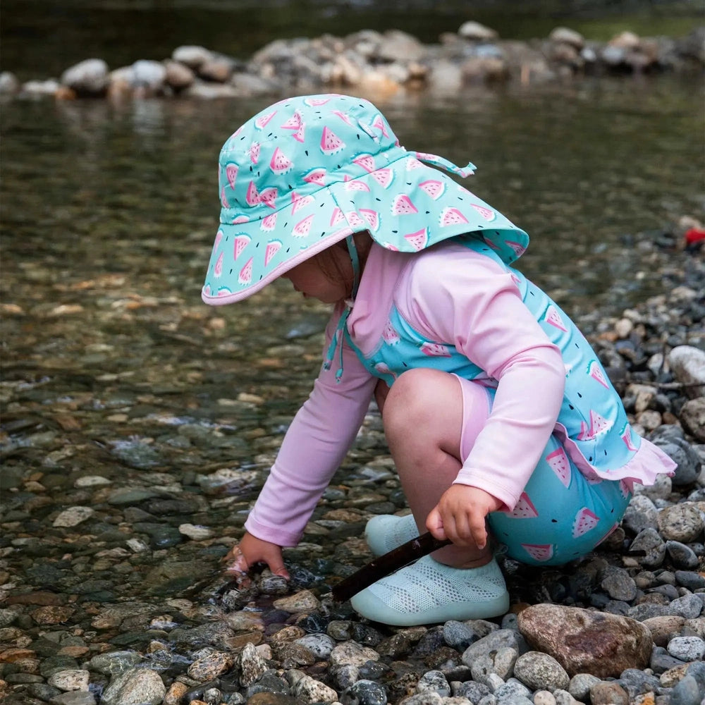 JAN & JUL Kids Water Repellent Adventure Hat, watermelon print in turquoise, as a child crouches by a rocky riverbank touching the water.