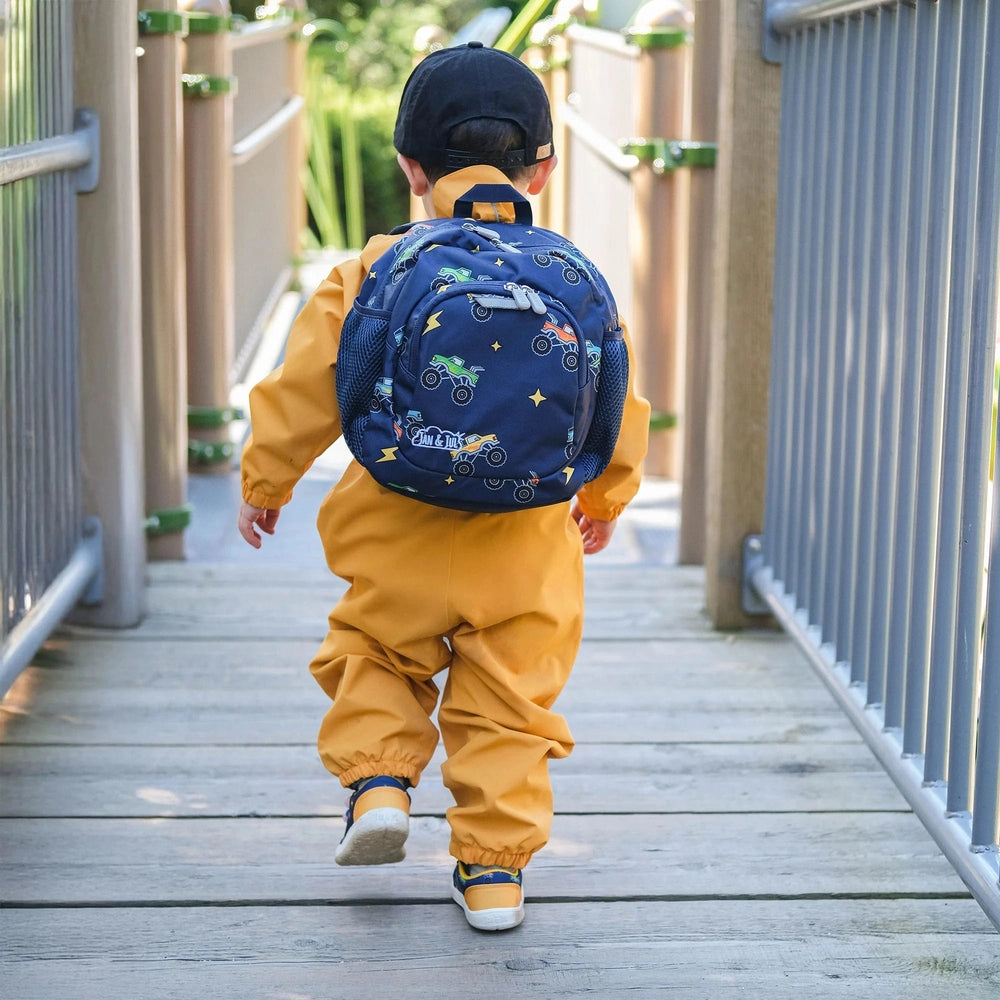 Jan & Jul Kids Mini Backpack in navy monster-truck print, worn by a child walking on a wooden bridge in mustard-yellow jumpsuit.