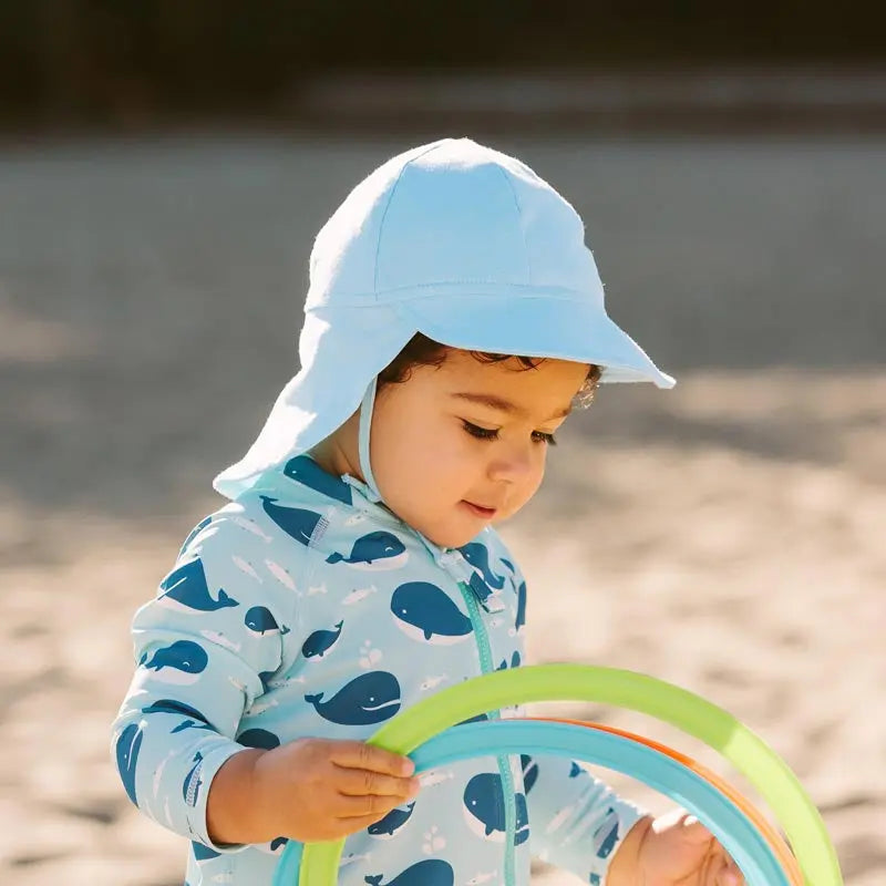 Jan & Jul Sun Soft Baby Cap in baby blue, a wide-brim sun hat with neck flap, worn by a toddler at the beach.