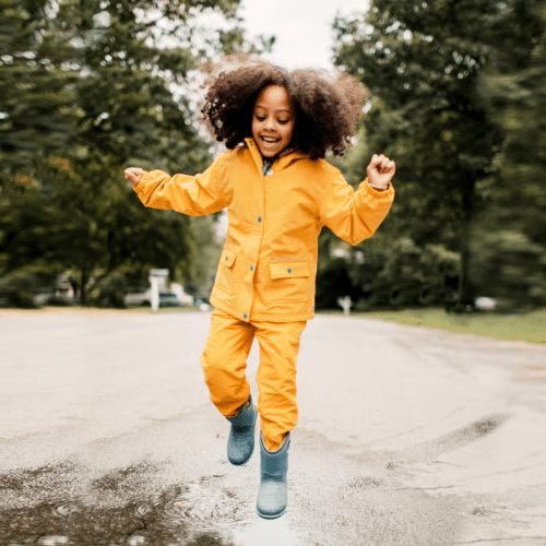 Jan & Jul Waterproof Cozy Dry Rain Pants Yellow worn by a child leaping over a puddle on a rainy street.