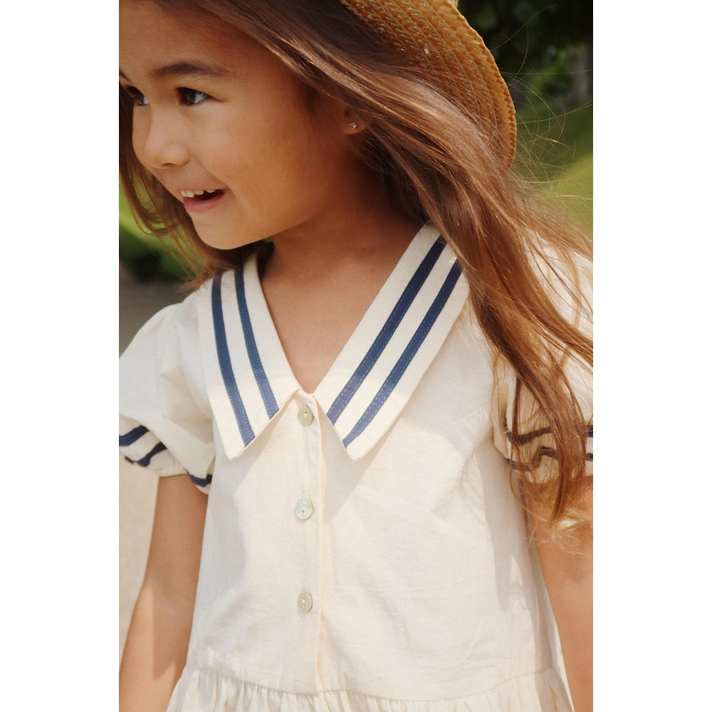 Konges Slojd Allie Sailor Dress GOTS Optic White; girl smiles in white dress with navy sailor collar and puff sleeves.