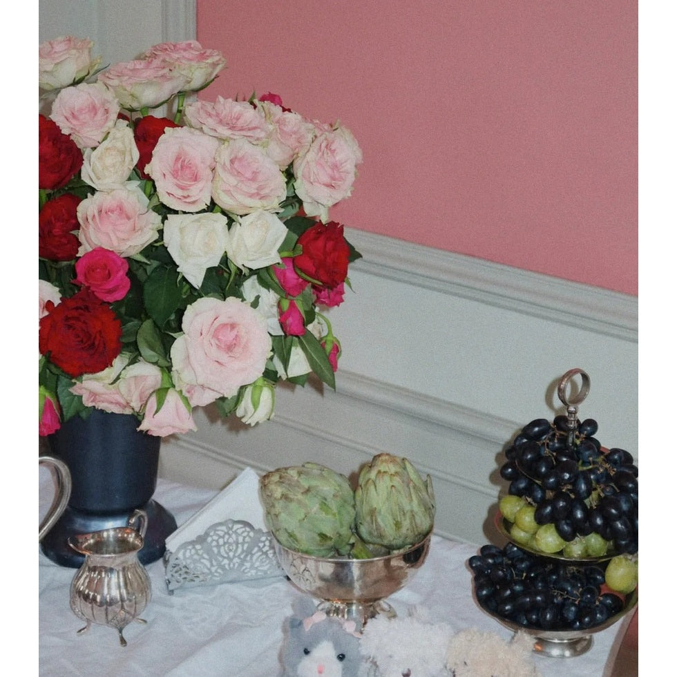 Konges Slojd Baby Animals plush toy sits on a lace table beside a pink rose bouquet and fruit stand.