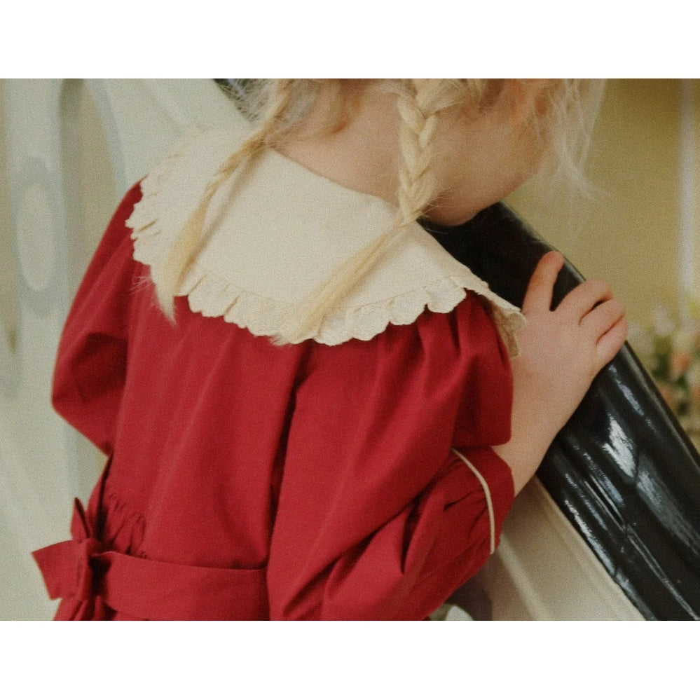 Konges Slojd Coeur Dress Red Dahlia worn by a girl with braided hair, holding a glossy black case, showcasing a cream lace collar and waist tie.