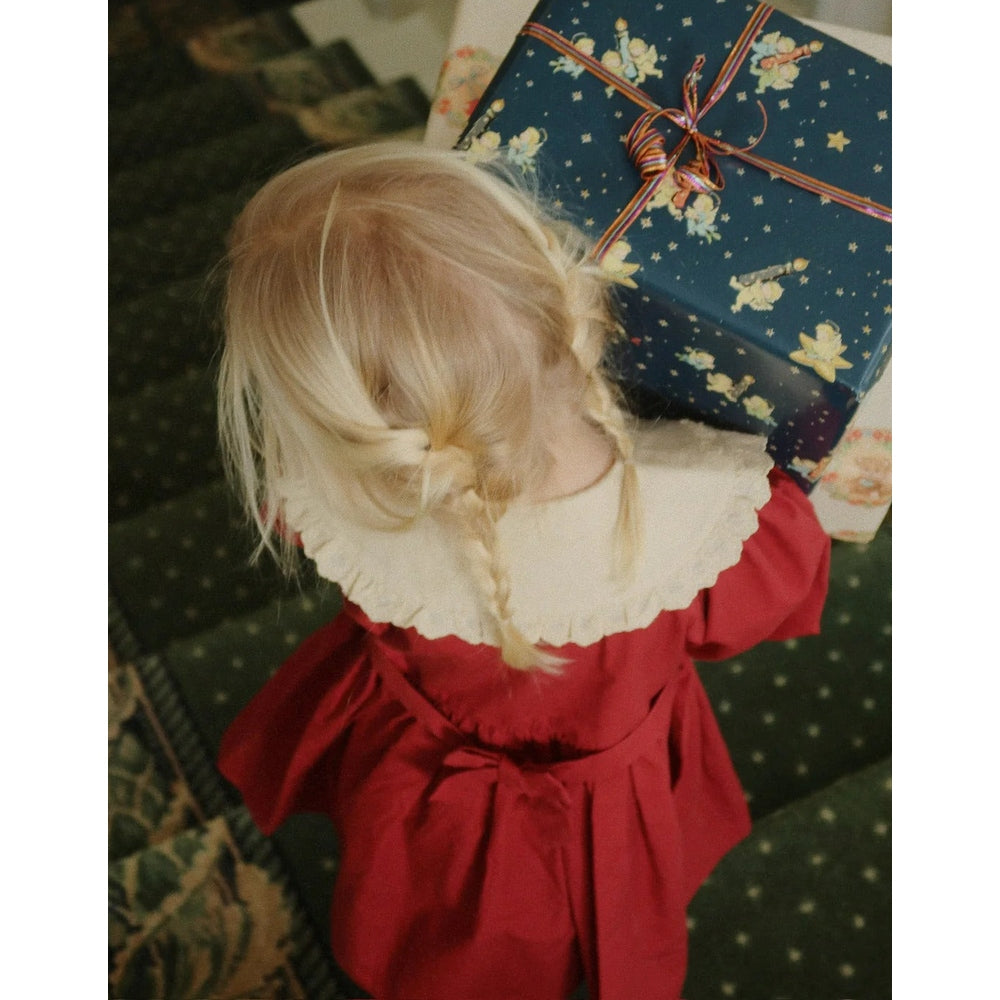 Konges Slojd Coeur Dress Red Dahlia on a toddler with a white scalloped collar, carrying a wrapped gift box.