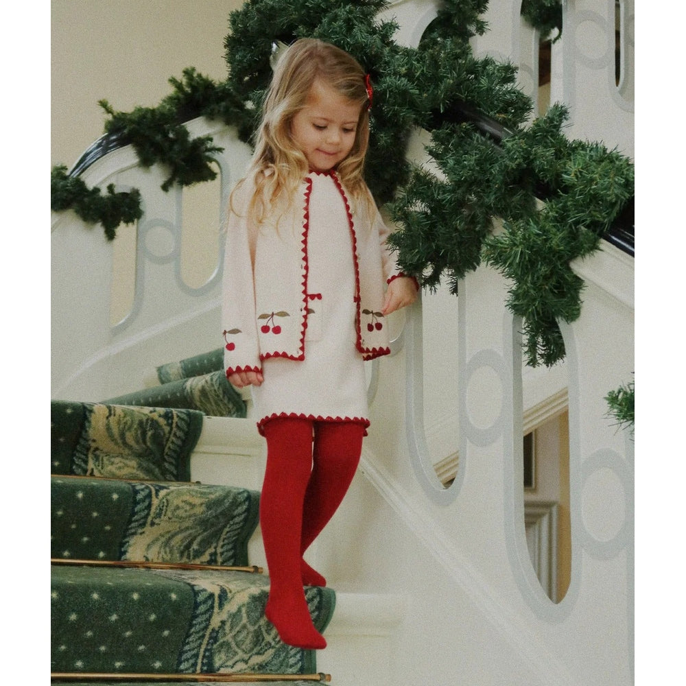 Konges Slojd Collette Cardigan Off White on a girl walking down a festively garlanded staircase in red tights.