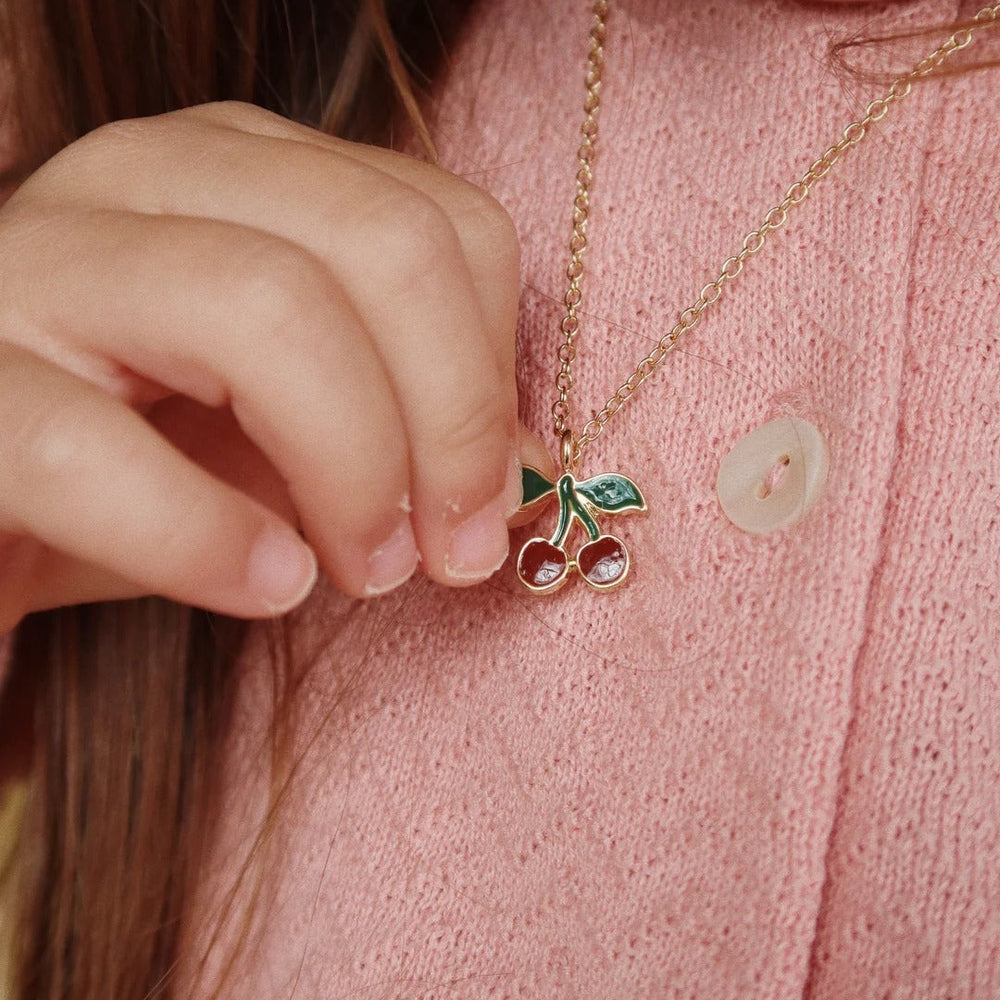 Konges Slojd Jewelry Set cherry pendant necklace with green leaves, held against pink knit cardigan.