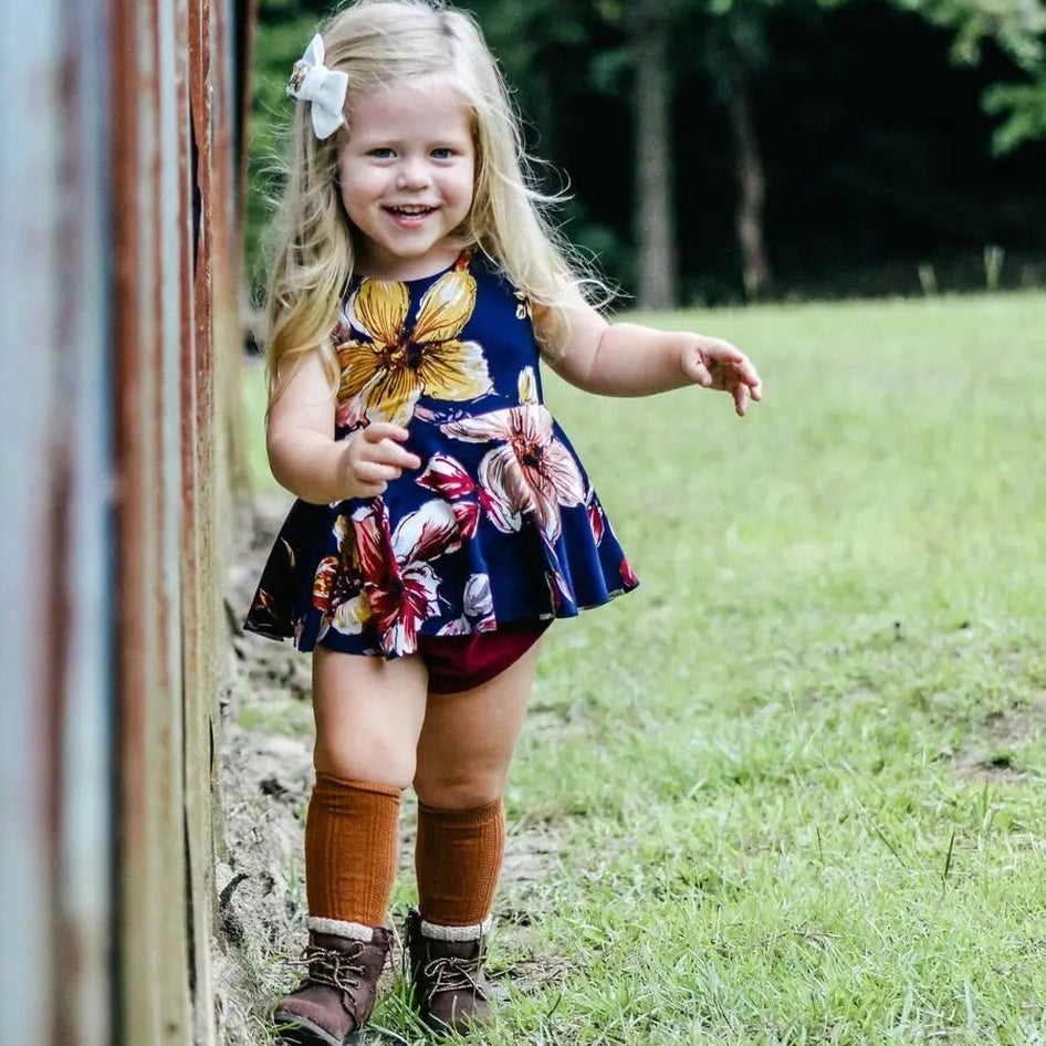 Little Stocking Co. Knee High Socks Almond on a smiling toddler walking along a fence in a floral dress.