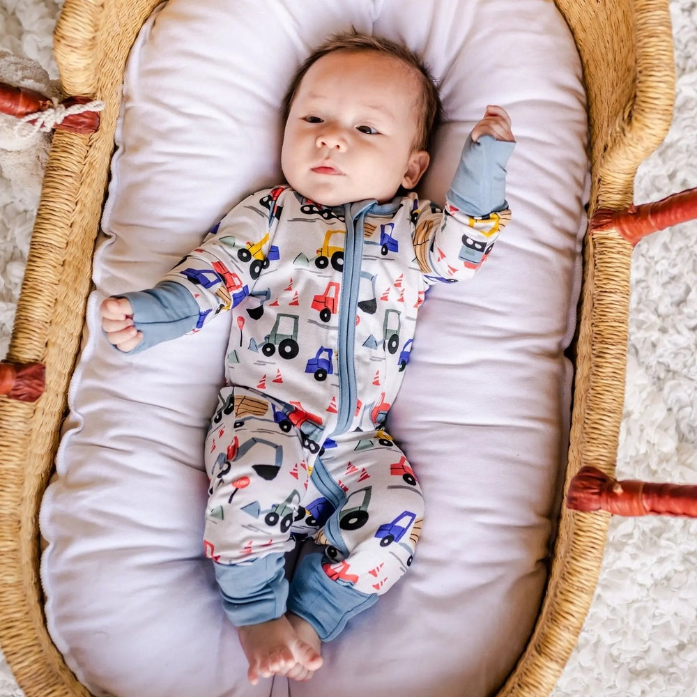 Littlesleepies Construction Bamboo Viscose Zippy sleeper; baby lies in a cushioned bassinet wearing a colorful truck-print outfit with arms up.