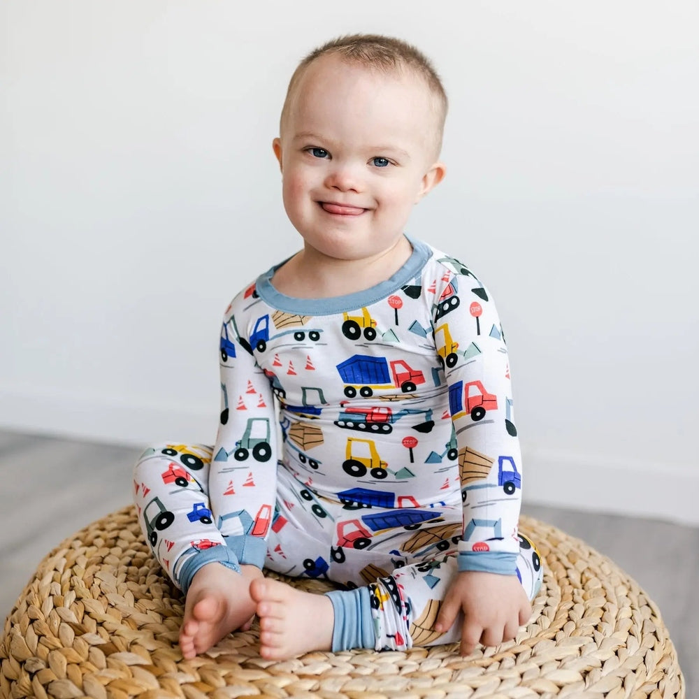 Littlesleepies Construction two-piece bamboo viscose pajama set with blue trim, baby sits cross-legged in truck-print pajamas.