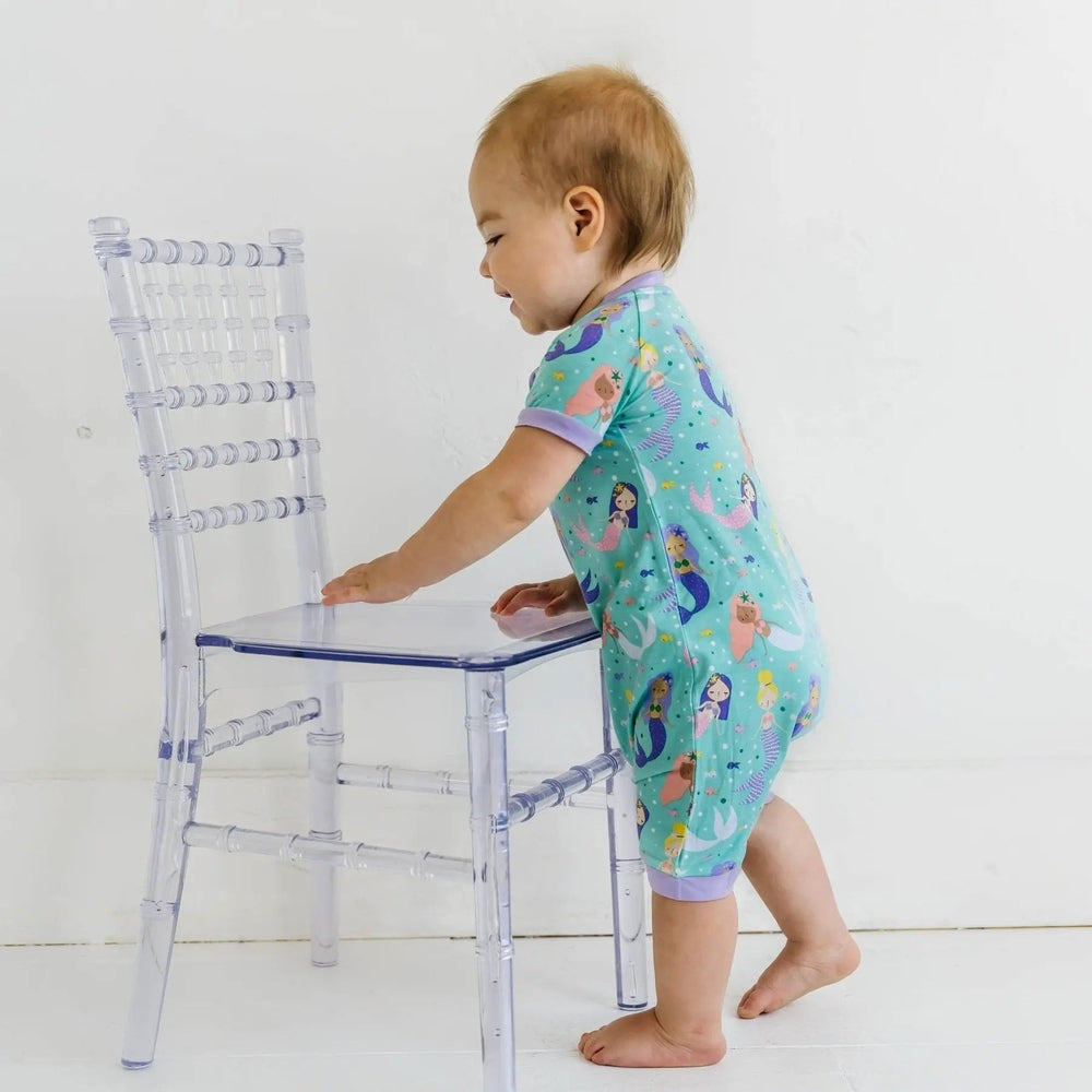 Littlesleepies Mermaid Magic Bamboo Viscose Shorty Zippy on a toddler standing by a clear chair in turquoise mermaid print.