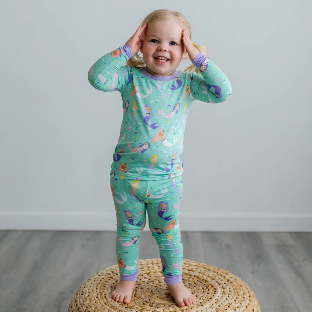 Littlesleepies Mermaid Magic Two Piece Bamboo Viscose Pajama Set worn by a smiling toddler in teal mermaid print, standing barefoot.