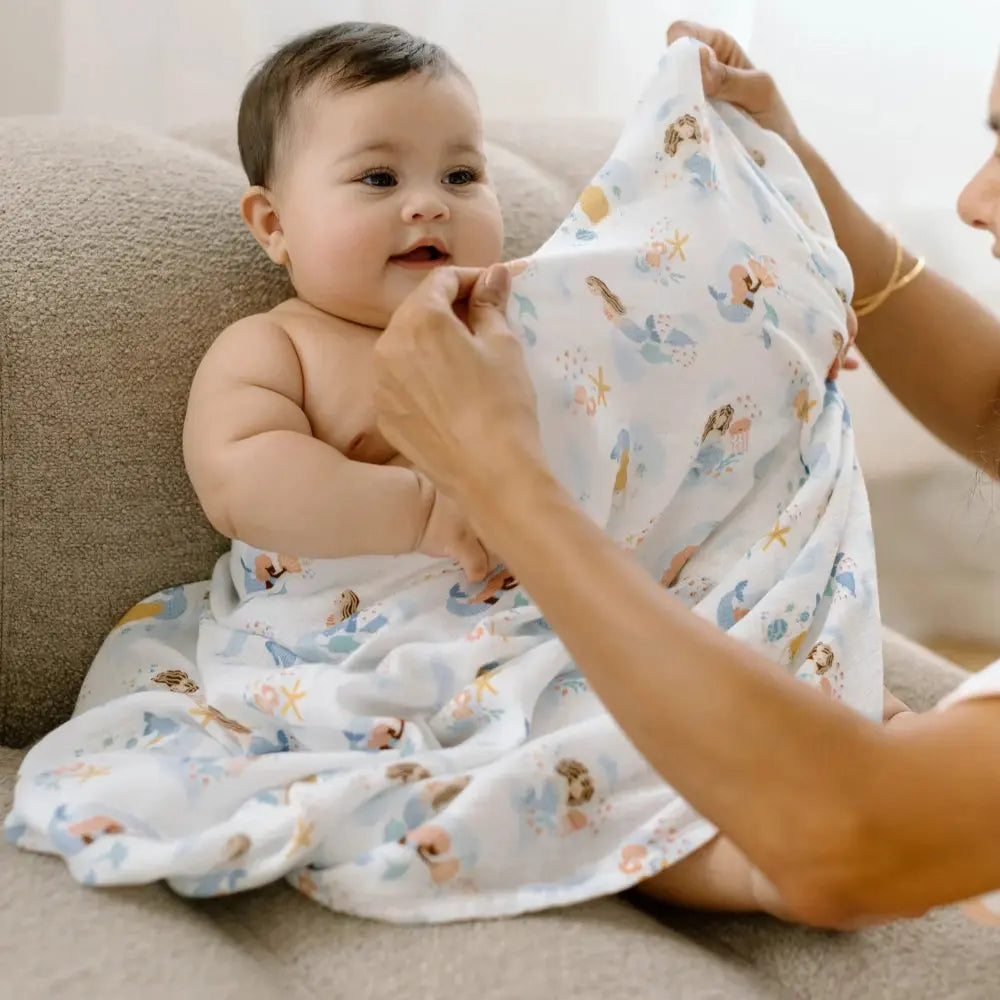 Loulou Lollipop Muslin Swaddle in light blue sea-print, shown snugly wrapped around baby by caregiver.