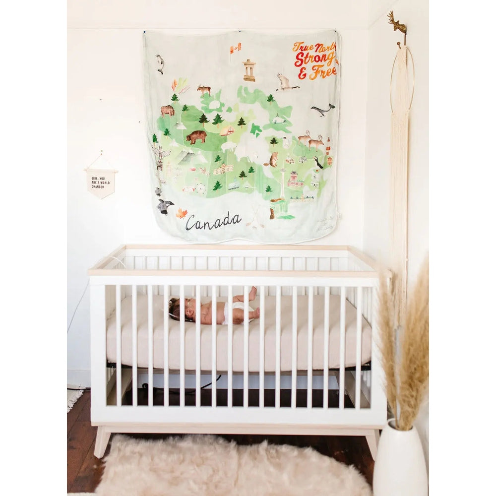 Loulou Lollipop Muslin Swaddle with Canada map print in green pastels, hung above a sleeping baby in a white crib.