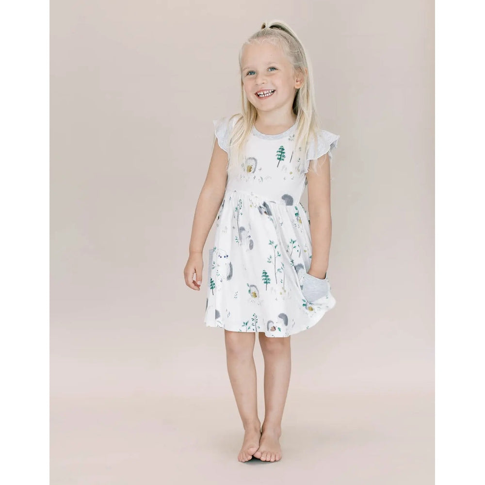 Loulou Lollipop Hedgehogs ruffle pocket dress in white cotton; girl stands barefoot, smiling, showcasing the woodland print.