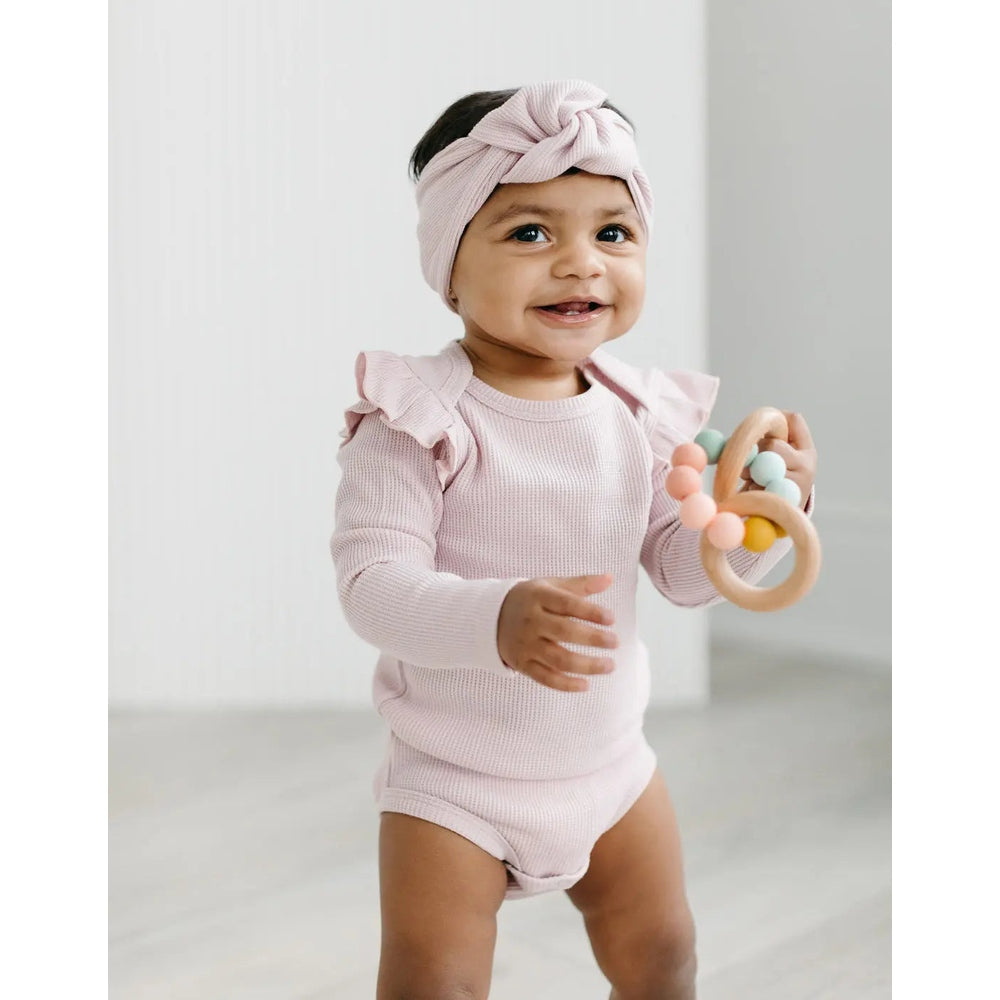 Loulou Lollipop Waffle Knotted Headband Blush Pink on a smiling baby in a matching knit outfit, knot front as she grips a pastel teether.