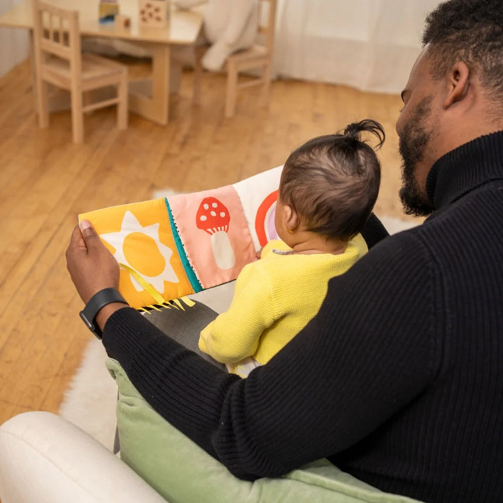 Manhattan Toy Wimmer Seek & Peek Book; infant in yellow sweater on dad's lap explores colorful fabric pages.