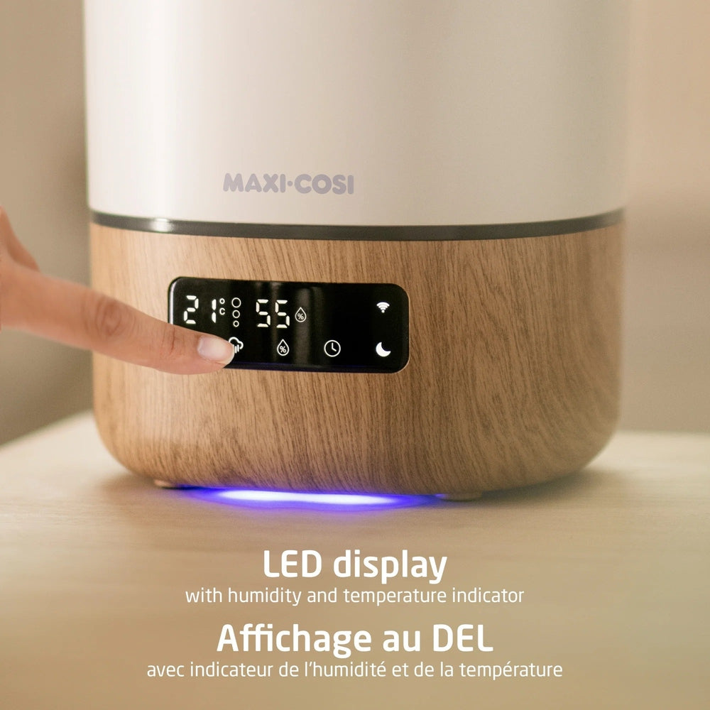 Maxi-Cosi Smart Humidifier with wood-grain base and LED display showing 21°C, 55% humidity as a finger taps the control.