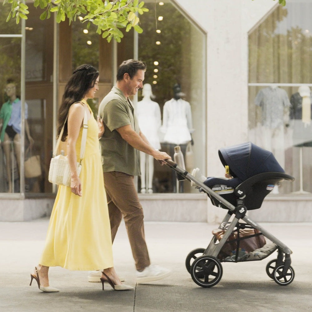 Maxi-Cosi Zelia Luxe 5-in-1 travel system stroller being pushed by a couple, navy canopy, bassinet attached.