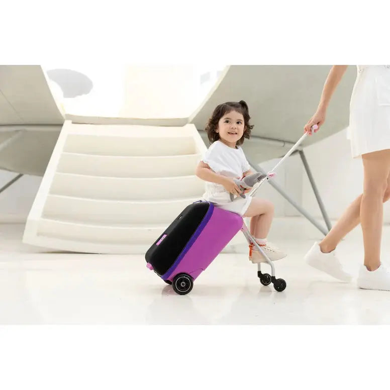 Micro Eazy Luggage purple kid’s suitcase with wheels, pulled by an adult while a child rides a scooter nearby.