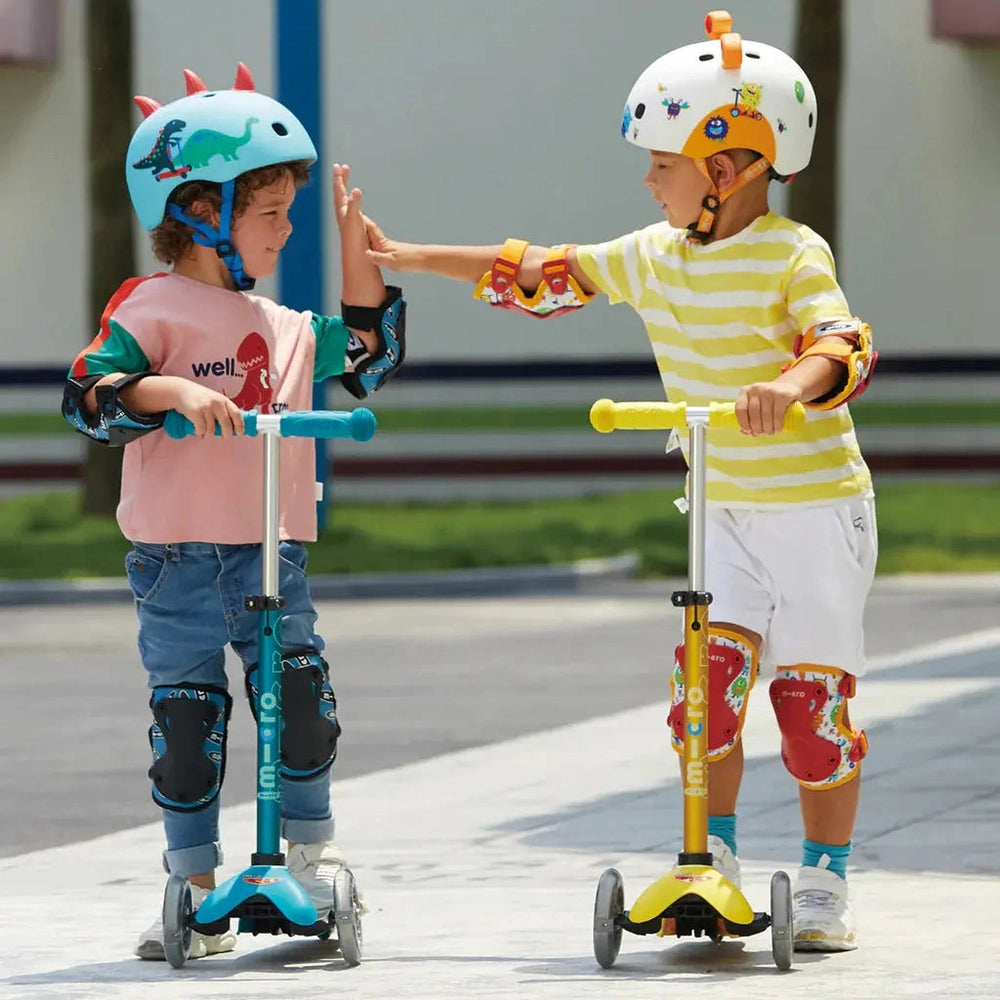 Micro Mini Deluxe LED Scooter pair; two teal blue and yellow scooters as kids high-five wearing helmets.
