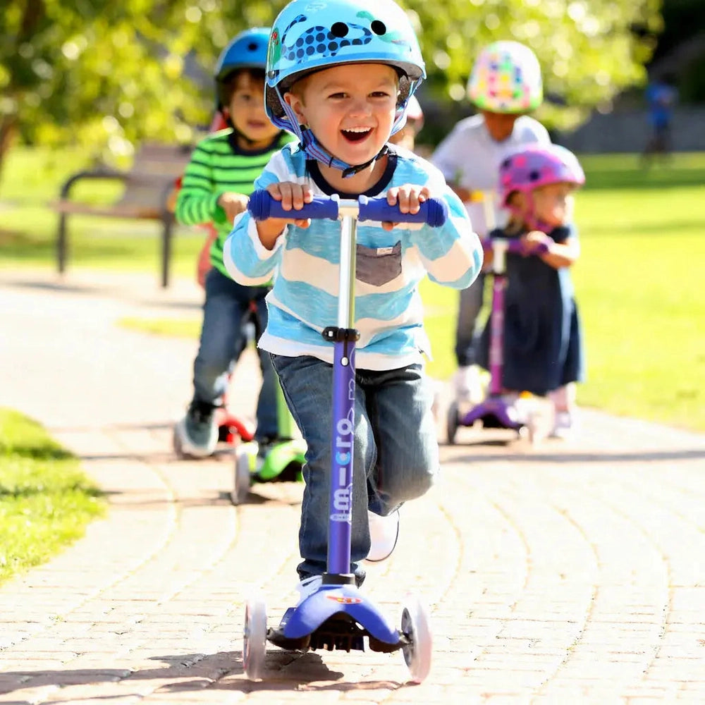 Micro Mini Deluxe Scooter with blue-purple frame; smiling helmeted child rides along a park path.