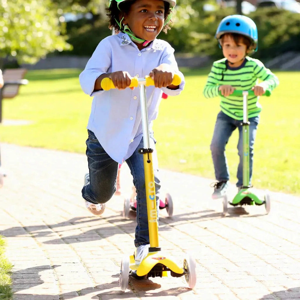 Micro Mini Deluxe Scooter, yellow three-wheel kid's scooter, child speeds along park path.