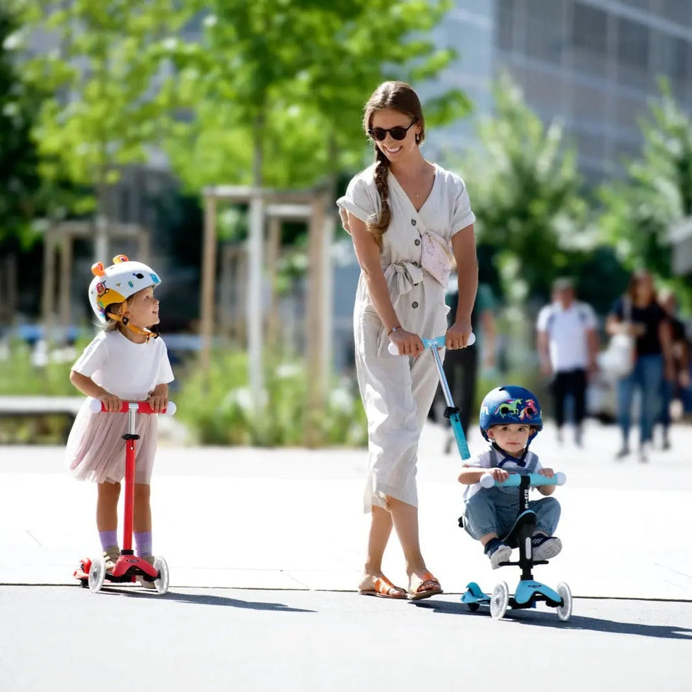 Micro Mini2Grow Deluxe LED scooter being ridden by two kids on red and blue scooters as mom supervises in a sunny park.