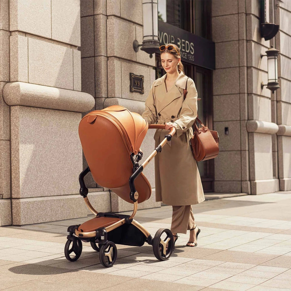 Mima Xari Max Stroller in tan leather, pushed by a woman in a trench coat along a city sidewalk.