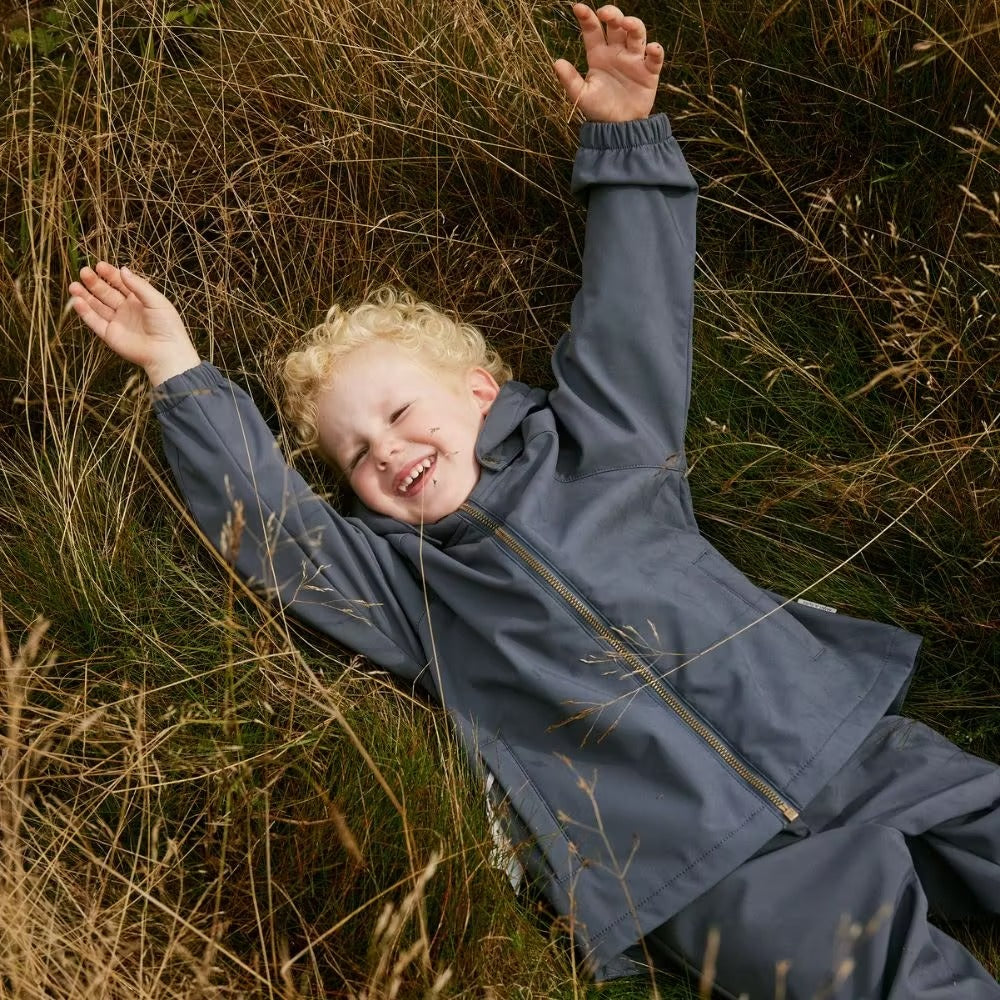 MINI A TURE Mataden Spring Softshell Jacket Ombre Blue worn by toddler lying in tall grass, front zipper visible.