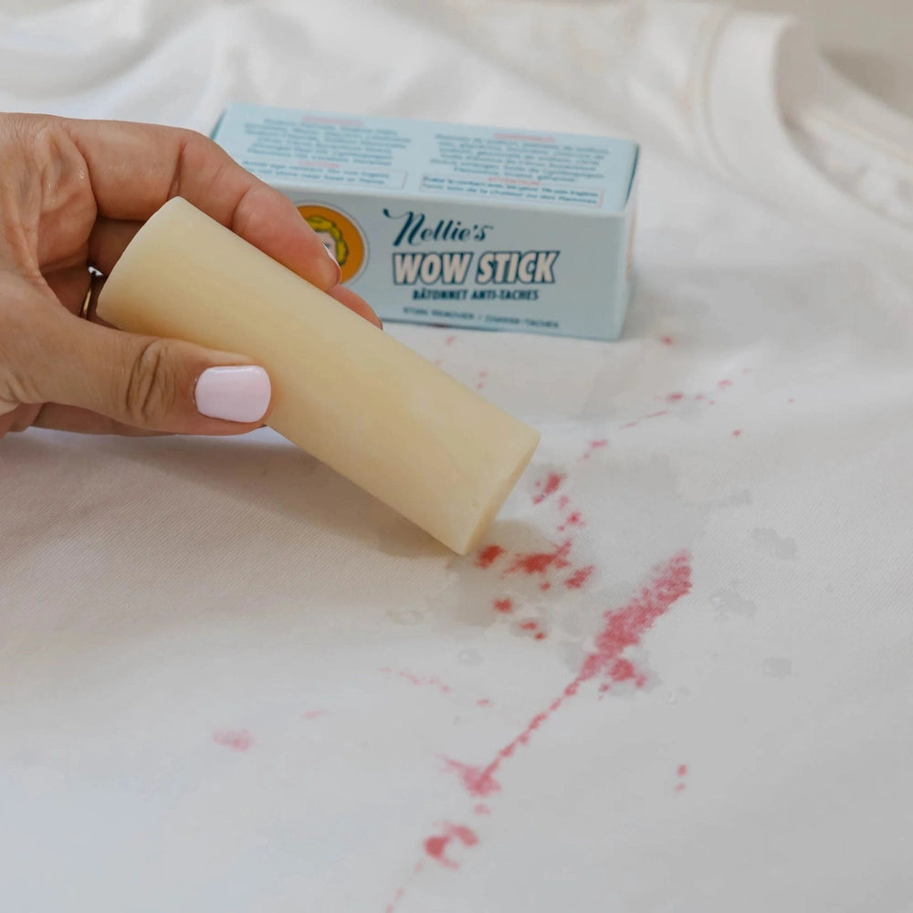 Nellie's Wow Stick Stain Remover bar, pale yellow, held by hand above a pink-stained white fabric.