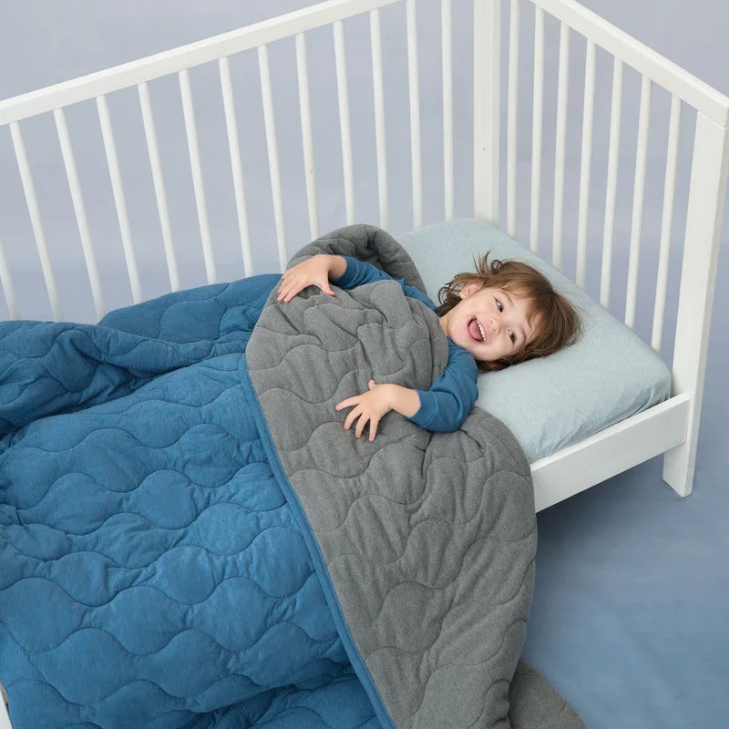Nest Designs Avocado Bamboo Cozy Blanket Medium Captain's Blue; blue quilted blanket tucked around a smiling child in a white crib.