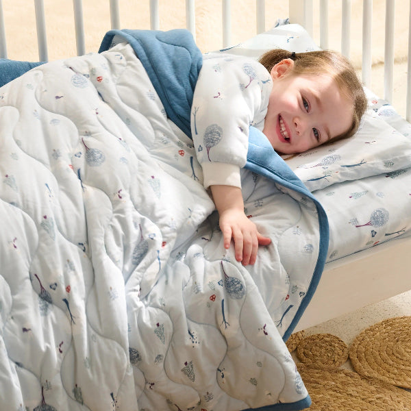 Nest Designs Bamboo Jersey Cozy Blanket Medium Snowy Snuggles wrapped around a smiling toddler in a cozy bed with blue trim.
