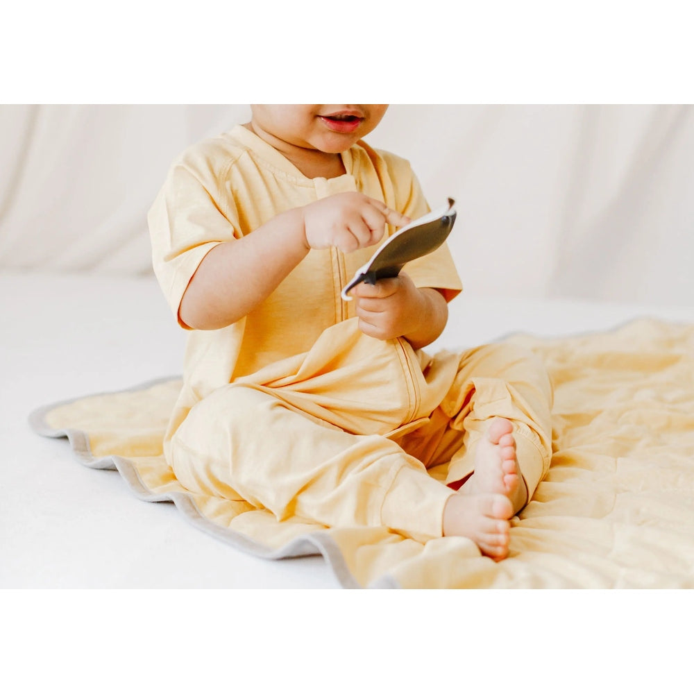 Nest Designs Bamboo Jersey Short Sleeve Footed Sleep Bag, 0.5 tog in Sunset Gold; baby sits on a cream blanket, holding the zipper open.